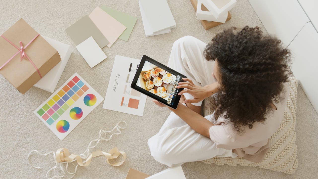 menu-02 Woman using tablet surrounded by design tools and gift box, showcasing creativity and online work.